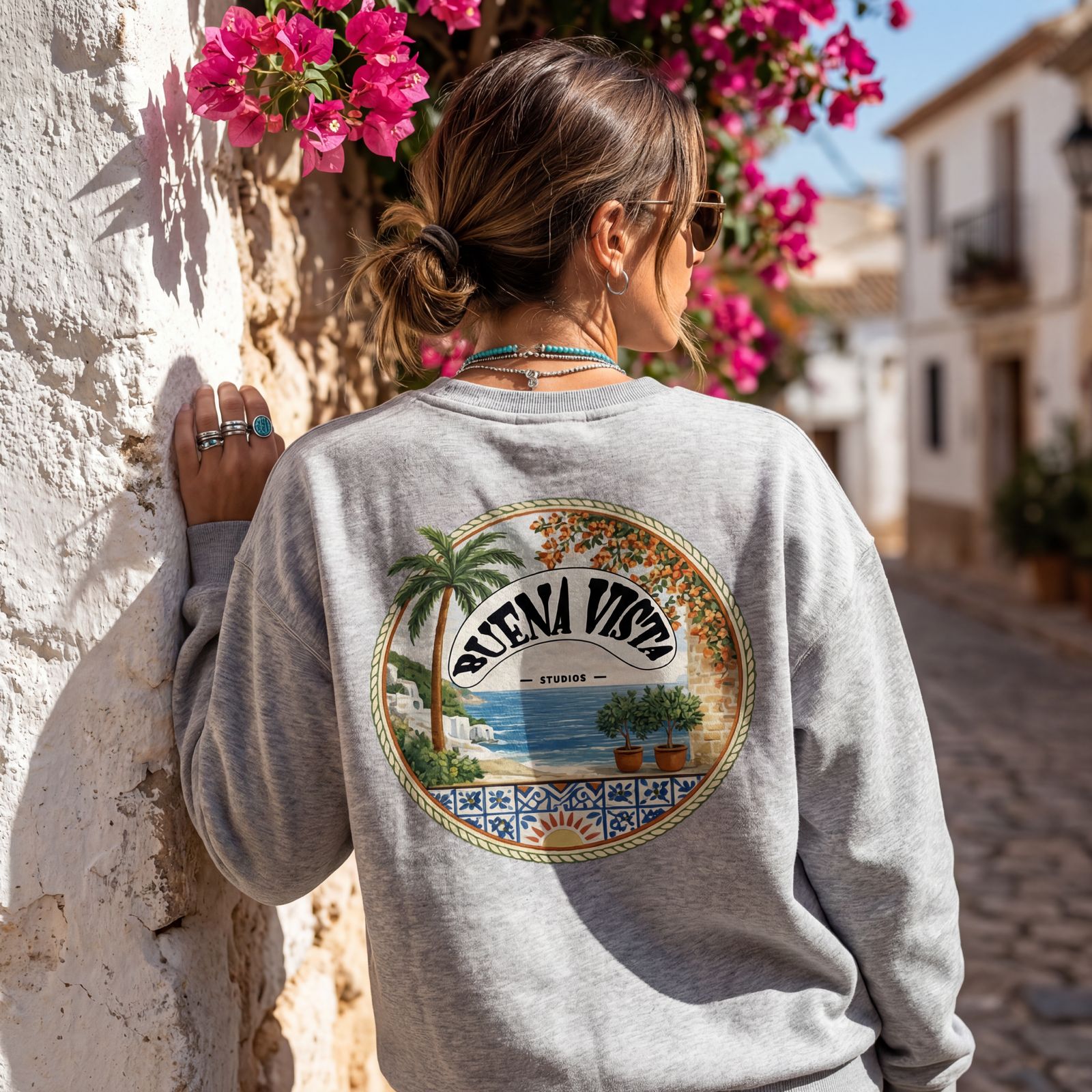 Woman wearing a gray sweatshirt with a tropical design, standing against a stone wall with pink flowers.